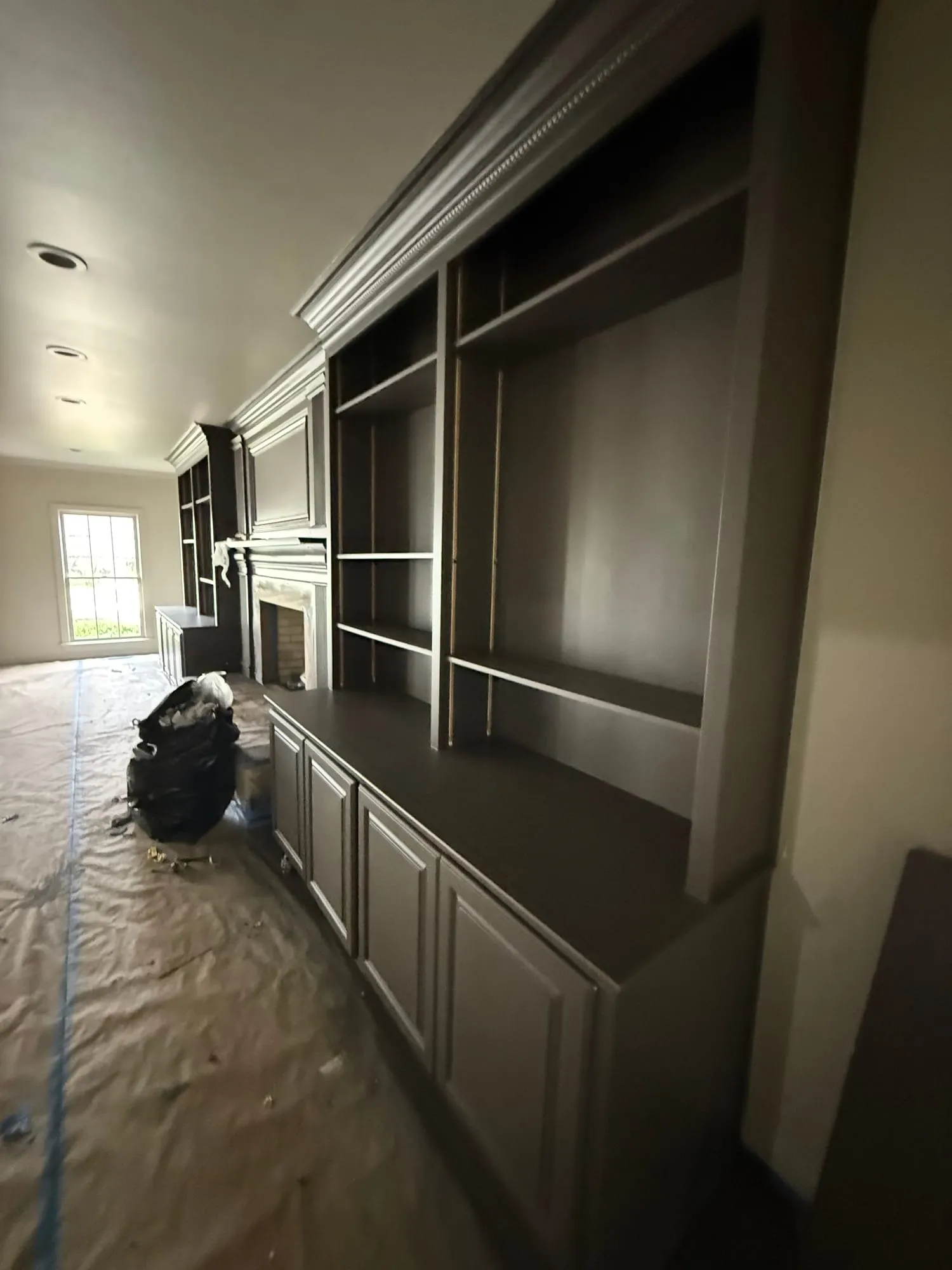 A floor-to-ceiling built-in wall unit with open shelving, base cabinets, and a fireplace mantel, all painted deep espresso against lighter walls, with drop cloths down mid-project.