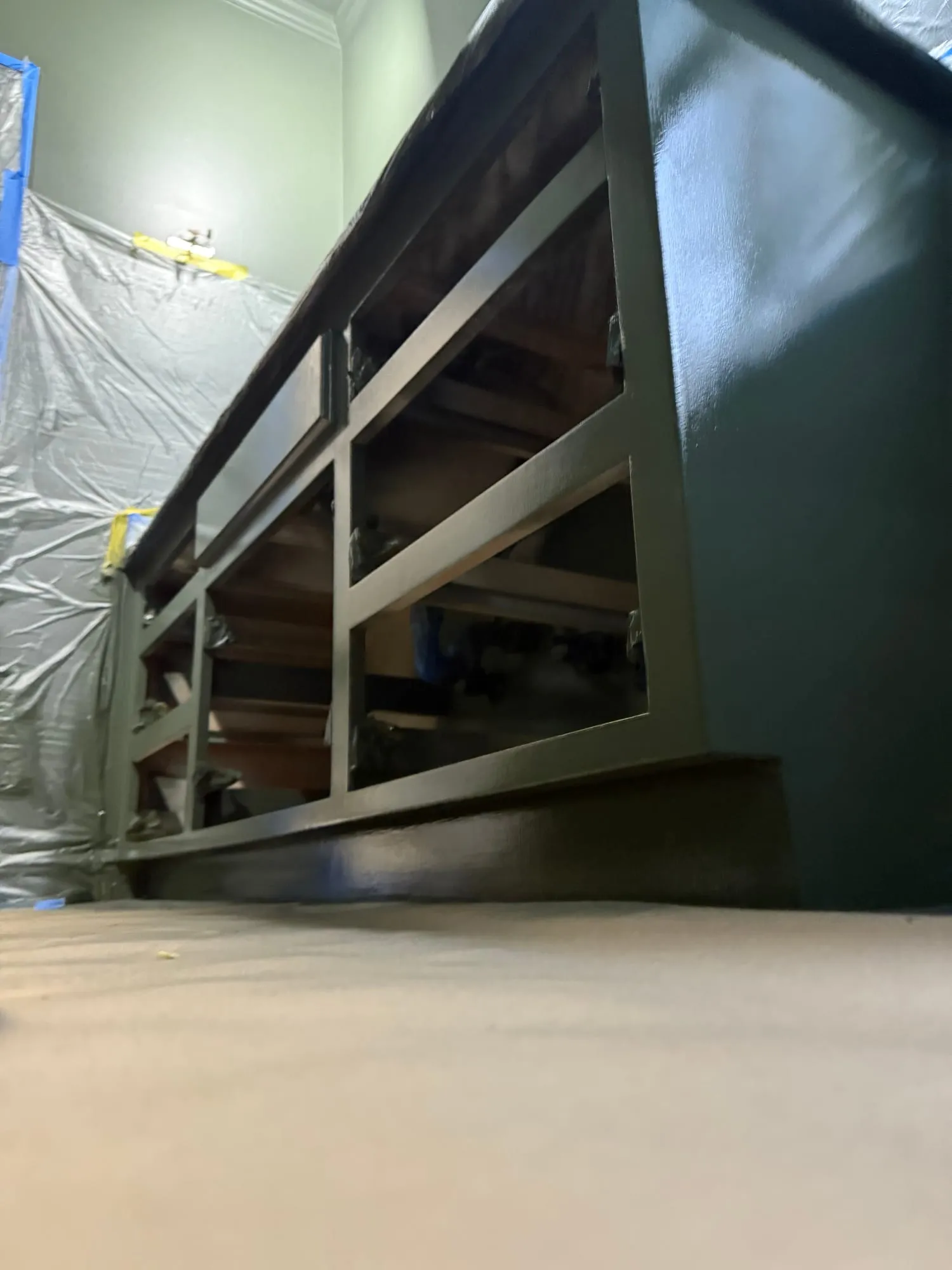A low built-in console cabinet finished in glossy dark green, doors removed, in a room masked off with plastic sheeting and sage walls behind.