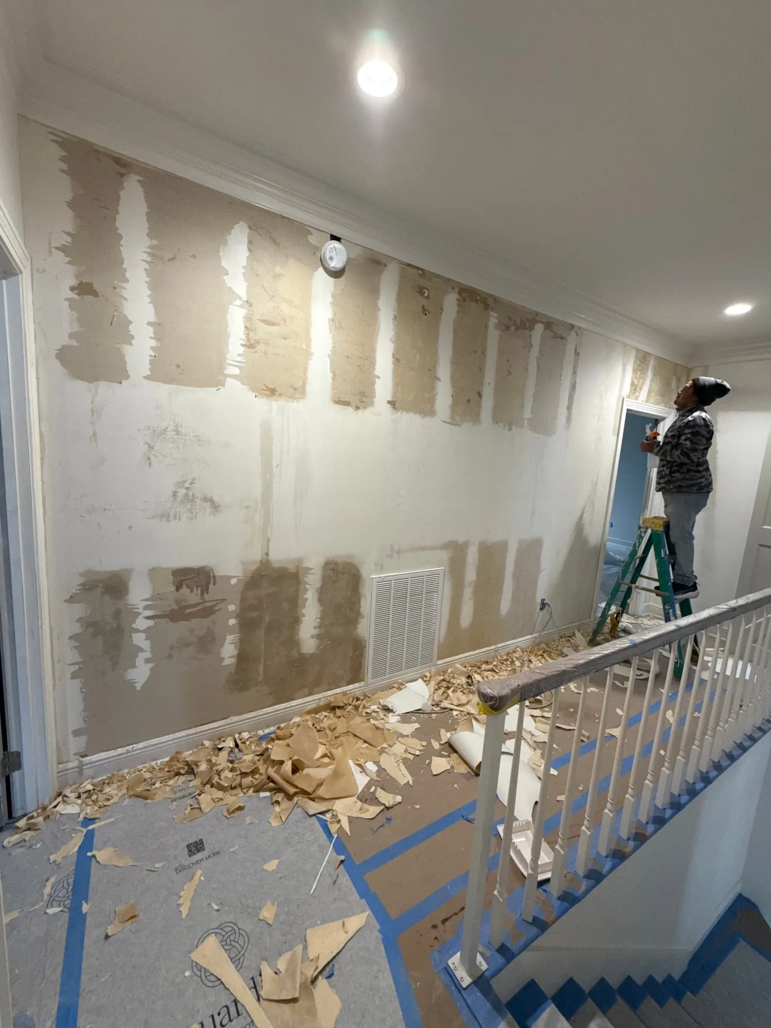 A worker on a ladder skim-coating a tall stairwell wall, old wallpaper stripped and piled on the covered floor, a painted handrail in the foreground.