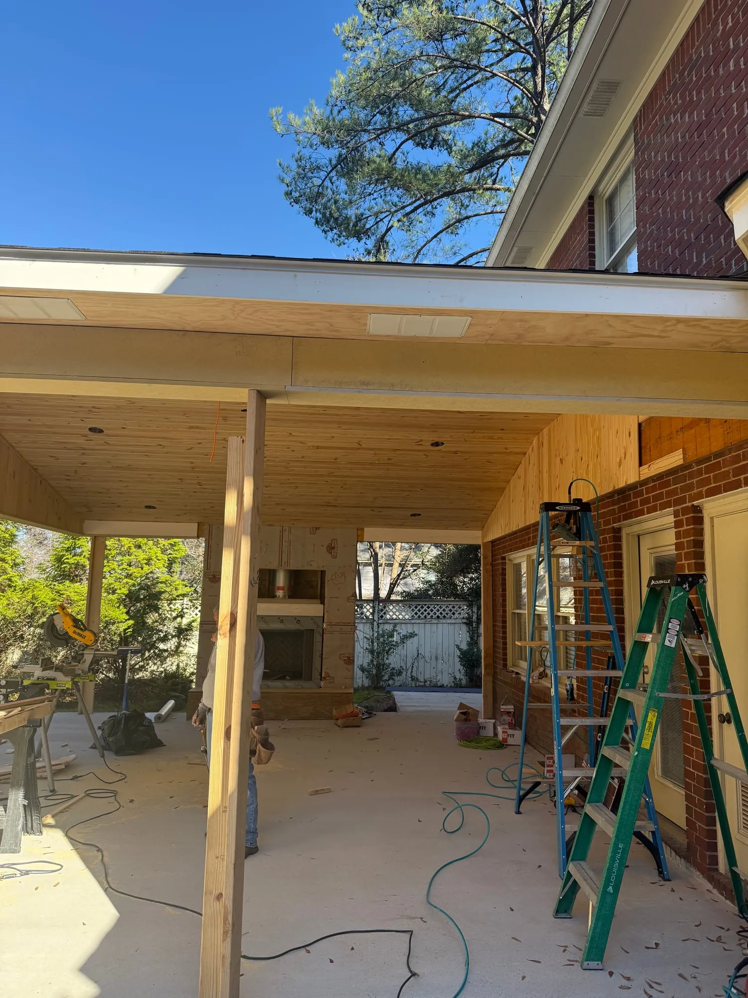 The same covered patio under construction, with a raw plywood ceiling, a framing post, ladders, and tools on the slab beside a half-built brick fireplace.